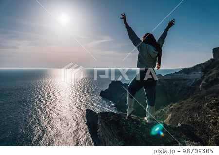 A young tourist Woman enjoying sunset over sea mountain landscape while sitting outdoor. Women's yoga fitness routine. Healthy lifestyle, harmony and meditation A young tourist Woman enjoying sunset over sea mountain landscape while sitting outdoor. Women's yoga fitness routine. Healthy lifestyle, harmony and meditation 98709305