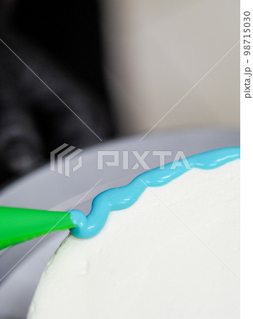hand pastry chef designer topping a frosted white cake with pastel light blue drip filling bag 98715030