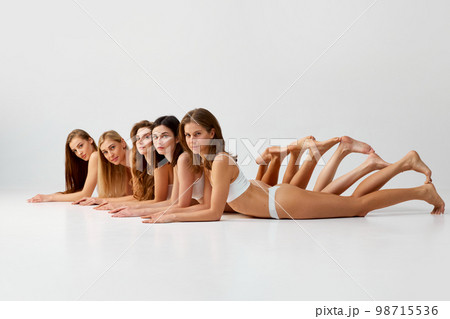 In a line. Group portrait of beautiful young women lying on floor, posing in white cotton underwear over grey studio background. Smooth skin. Concept of natural beauty, body care In a line. Group portrait of beautiful young women lying on floor, posing in white cotton underwear over grey studio background. Smooth skin. Concept of natural beauty, body care 98715536