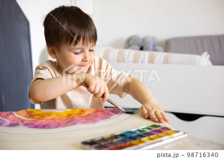 Confident cute male kid art drawing multicolored rainbow arch use paints and brush on table 98716340