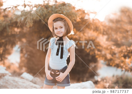 Smiling child girl 4-5 year old wearing straw hat with laces, swimsuit and denim shorts outdoors. Looking at camera. Summer time. Smiling child girl 4-5 year old wearing straw hat with laces, swimsuit and denim shorts outdoors. Looking at camera. Summer time. 98717737