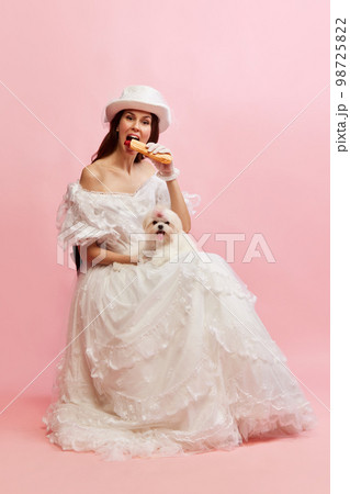 Eating hotdog. Portrait of beautiful lady in white vintage dress posing with dog over pink background. Concept of 19th century, fashion, comparison of eras 98725822