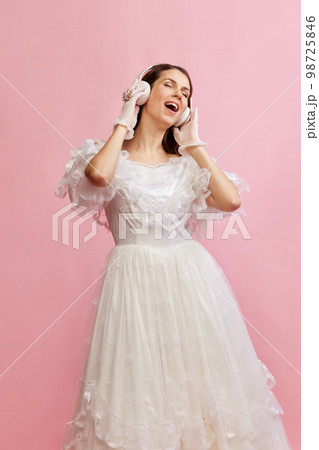 Portrait of beautiful lady in white vintage dress posing, listening to music in headphones over pink background. Concept of 19th century, fashion, comparison of eras Portrait of beautiful lady in white vintage dress posing, listening to music in headphones over pink background. Concept of 19th century, fashion, comparison of eras 98725846