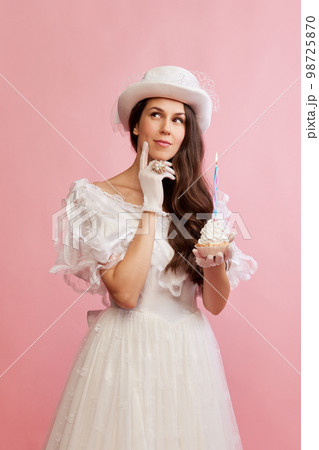Making wishes. Portrait of beautiful lady in white vintage dress posing with birthday cupcake over pink background. Concept of 19th century, fashion, comparison of eras 98725870