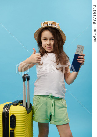 Cute little girl in summer wear, shows thumb up at camera, poses with flight ticket and yellow suitcase, blue background 98726143