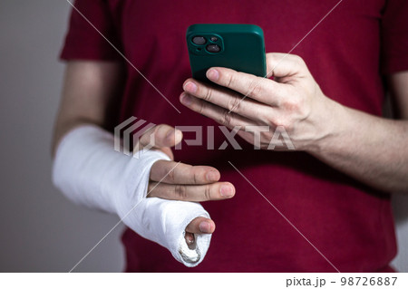 A man in a red T-shirt with a broken arm in a cast holds and uses a phone Close-up A man in a red T-shirt with a broken arm in a cast holds and uses a phone Close-up 98726887