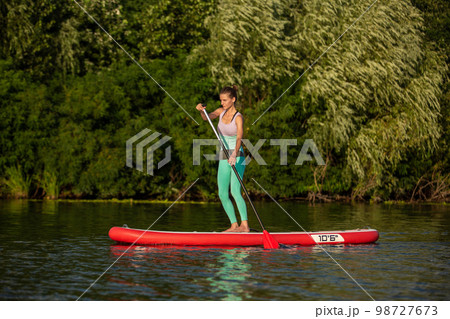 Young athletic woman doing fitness on a board with an oar on a lake. 98727673