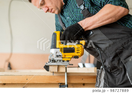 A man cuts a wooden plank with an electric jigsaw in a workshop. 98727889