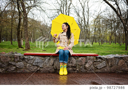Coping with stress and anxiety. Practicing mindfulness and relaxation techniques. Happy senior woman in yellow rain coat with yellow umbrella walking and enjoying life in rainy park 98727986