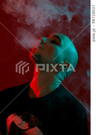 Close-up studio shot of a young bald guy vaping, blowing out a cloud of smoke on red background. 98728037