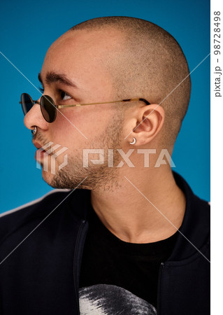 Studio shot of a young tattoed bald man posing against a blue background. 90s style. 98728498