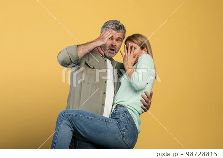 Portrait of scared middle aged couple covering mouth and eyes, woman jumping on man, looking at camera Portrait of scared middle aged couple covering mouth and eyes, woman jumping on man, looking at camera 98728815