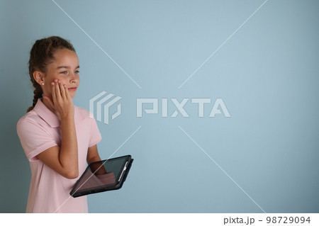 Little emotional teenage girl in pink shirt 11, 12 years old on an isolated blue background with a tablet in her hands.Children's studio portrait. Place the text to copy the place for the inscription Little emotional teenage girl in pink shirt 11, 12 years old on an isolated blue background with a tablet in her hands.Children's studio portrait. Place the text to copy the place for the inscription 98729094