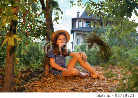 Country summer. Cute caucasian little girl wearing straw hat sits on the ground in garden. The concept of local farming and psychology 98729321