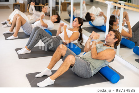 Middle-aged man practicing pilates with roller on gray mat in gym room Middle-aged man practicing pilates with roller on gray mat in gym room 98734938