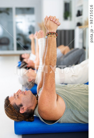 Middle-aged man practicing pilates with roller on gray mat in gym room 98735016