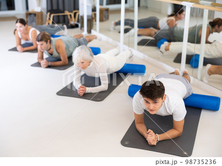 Positive athletic male performing set of exercises with pilates roller during group class in fitness studio. 98735236