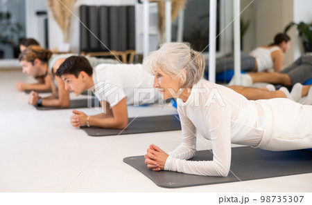Active senior woman performs exercises with a pilates roller while lying on a mat in the hall of a modern fitness studio. 98735307