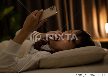 ベッド・寝室であくびをしながらスマホを操作する女性 98736043