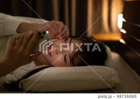 ベッド・寝室で目を擦りながらスマホを操作する女性 98736089