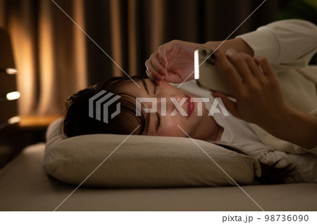 ベッド・寝室で目を擦りながらスマホを操作する女性 98736090