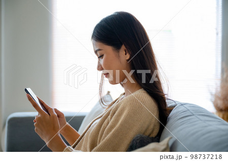 Happy young asian woman relax on comfortable couch at home texting messaging on smartphone 98737218