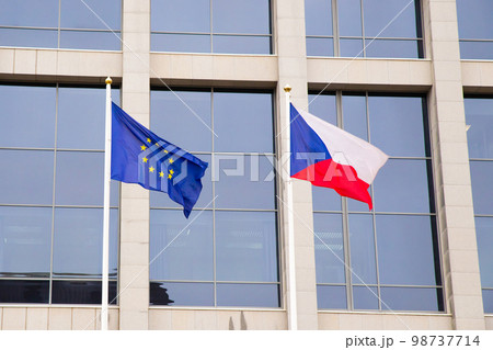 EU flag and Czech Republic flag on flagpole on modern consulate building. Flag of European Union and Czech flag on glass blue facade. 98737714