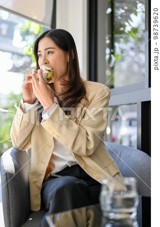 Hungry Asian businesswoman having an easy breakfast at the cafe, eating a healthy sandwich. Hungry Asian businesswoman having an easy breakfast at the cafe, eating a healthy sandwich. 98739620