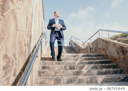 Confident middle age businessman with briefcase walking downstairs. Close-up of businessman wearing blue suit holding bag and coffee cup walking down the stairs 98742649