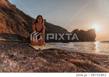Young woman with long hair in black swimsuit and boho style braclets practicing outdoors on yoga mat by the sea on a sunset. Women's yoga fitness routine. Healthy lifestyle, harmony and meditation 98742863