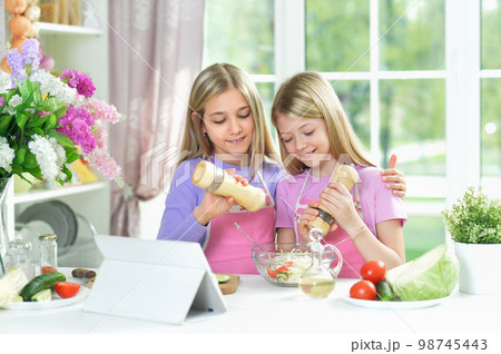 Two girls in pink aprons preparing salad on kitchen with tablet 98745443