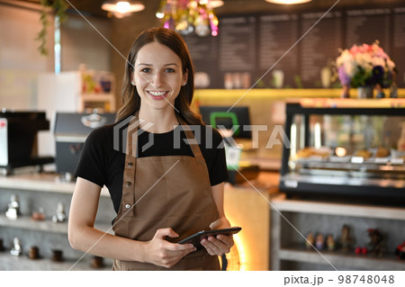 Half length of cheerful caucasian entrepreneur in apron standing in modern cafe interior and using digital tablet Half length of cheerful caucasian entrepreneur in apron standing in modern cafe interior and using digital tablet 98748048