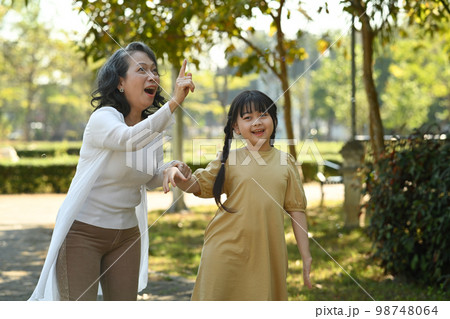 Joyful little girl and grandmother walking in public park surrounded by green trees at sunlight morning. Family, generation concept Joyful little girl and grandmother walking in public park surrounded by green trees at sunlight morning. Family, generation concept 98748064