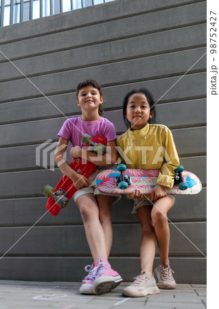 Happy friends posing together in city, standing in front of concrete wall, holding skateboards and looking at camera. 98752427