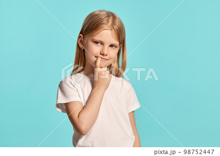 Close-up studio shot of a lovely blonde little girl in a white t-shirt posing against a blue background. 98752447