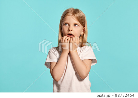 Close-up studio shot of a lovely blonde little girl in a white t-shirt posing against a blue background. 98752454