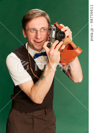 Amateur photographer. Portrait of man in stylish classical clothes, shorts and vest posing over green studio background. Concept of emotions, facial expression, lifestyle 98756611