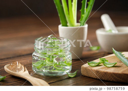 Glass jar of aloe vera gel, sliced aloe plant on a cutting board. Mortars of aloe vera leaves on background. Natural cosmetic and healing remedy. Glass jar of aloe vera gel, sliced aloe plant on a cutting board. Mortars of aloe vera leaves on background. Natural cosmetic and healing remedy. 98758759