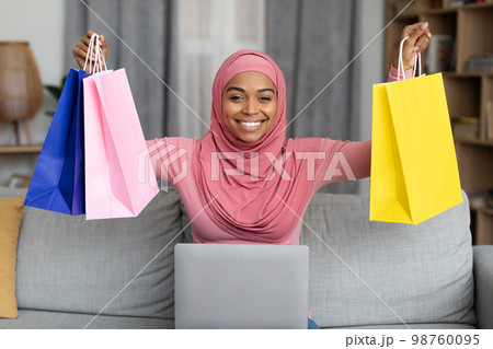 Excited black lady in hijab sitting on sofa with laptop and lifting gift bags above her head, happy to get big discount 98760095