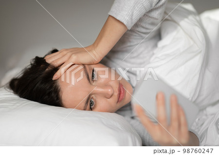 Sad unhappy young european woman lies on white bed, wakes up, reads message on smartphone in bedroom 98760247