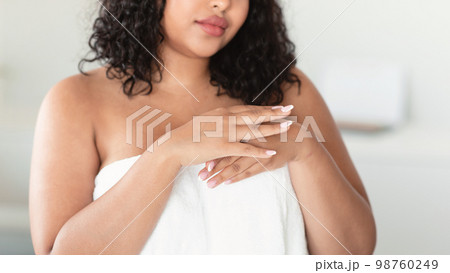 Unrecognizable plus size lady in white towel applying moisturising lotion or nourishing cream on hands, home interior 98760249