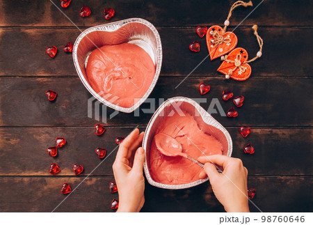Women's hands with spoon put the dough in form of foil for baking cake in form of heart. 98760646