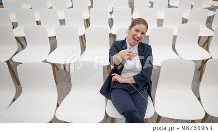 Red-haired caucasian business woman sits on the front row in an empty conference room and points her finger at the camera.  98761953