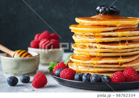 Delicious pancakes, with fresh blueberries, raspberry and maple syrup or honey on a dark background Delicious pancakes, with fresh blueberries, raspberry and maple syrup or honey on a dark background 98762334