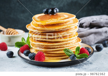 Delicious pancakes, with fresh blueberries, raspberry and maple syrup or honey on a dark background Delicious pancakes, with fresh blueberries, raspberry and maple syrup or honey on a dark background 98762362
