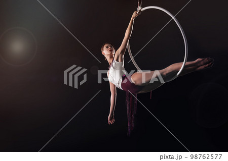 Young woman performing acrobatic element on aerial ring against dark background Young woman performing acrobatic element on aerial ring against dark background 98762577