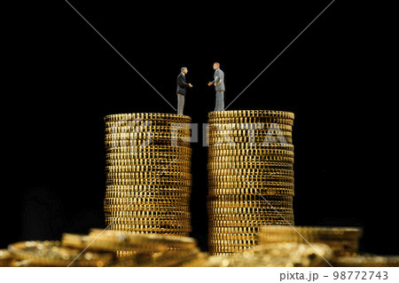高く積み重ねたコインの上で握手をするミニチュアのビジネスパーソン 98772743