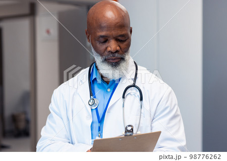 African american senior male doctor reading notes in hospital corridor, copy space 98776262