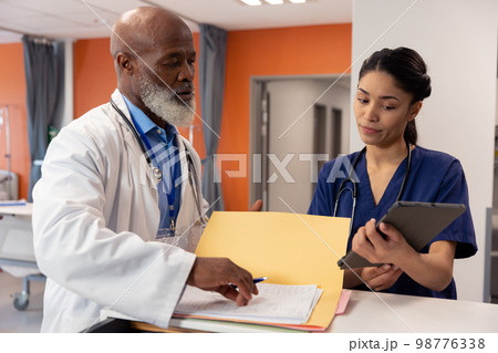 Diverse female and senior male doctor using tablet and talking in hospital ward, copy space 98776338