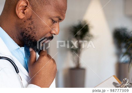 Thoughtful african american male doctor reading notes on clipboard in hospital corridor Thoughtful african american male doctor reading notes on clipboard in hospital corridor 98777309
